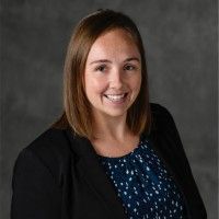 Woman with light brown hair smiles, wearing a black blazer over a blue and white patterned top. Gray background.