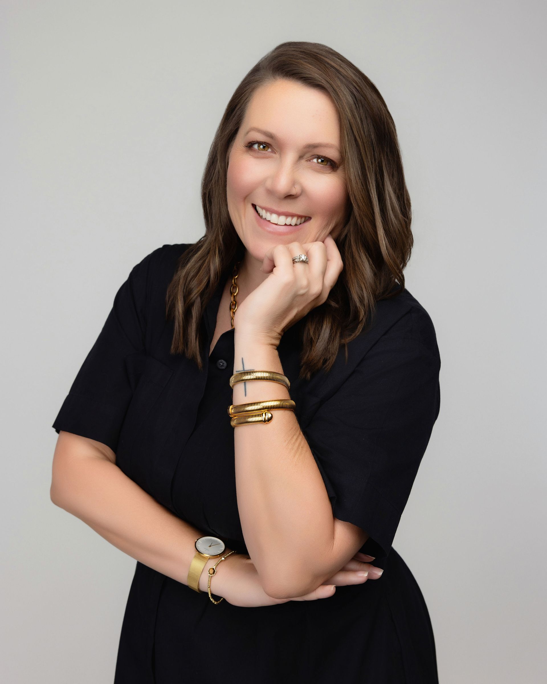 Woman in black dress smiles, hand to her chin, gold bracelets and watch, against a gray background.