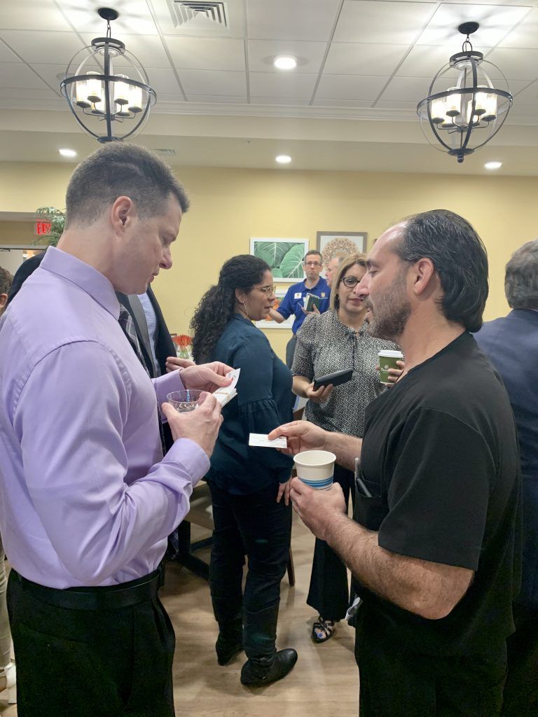 Two men in a room talking, both holding small papers. Several other people are in the background.