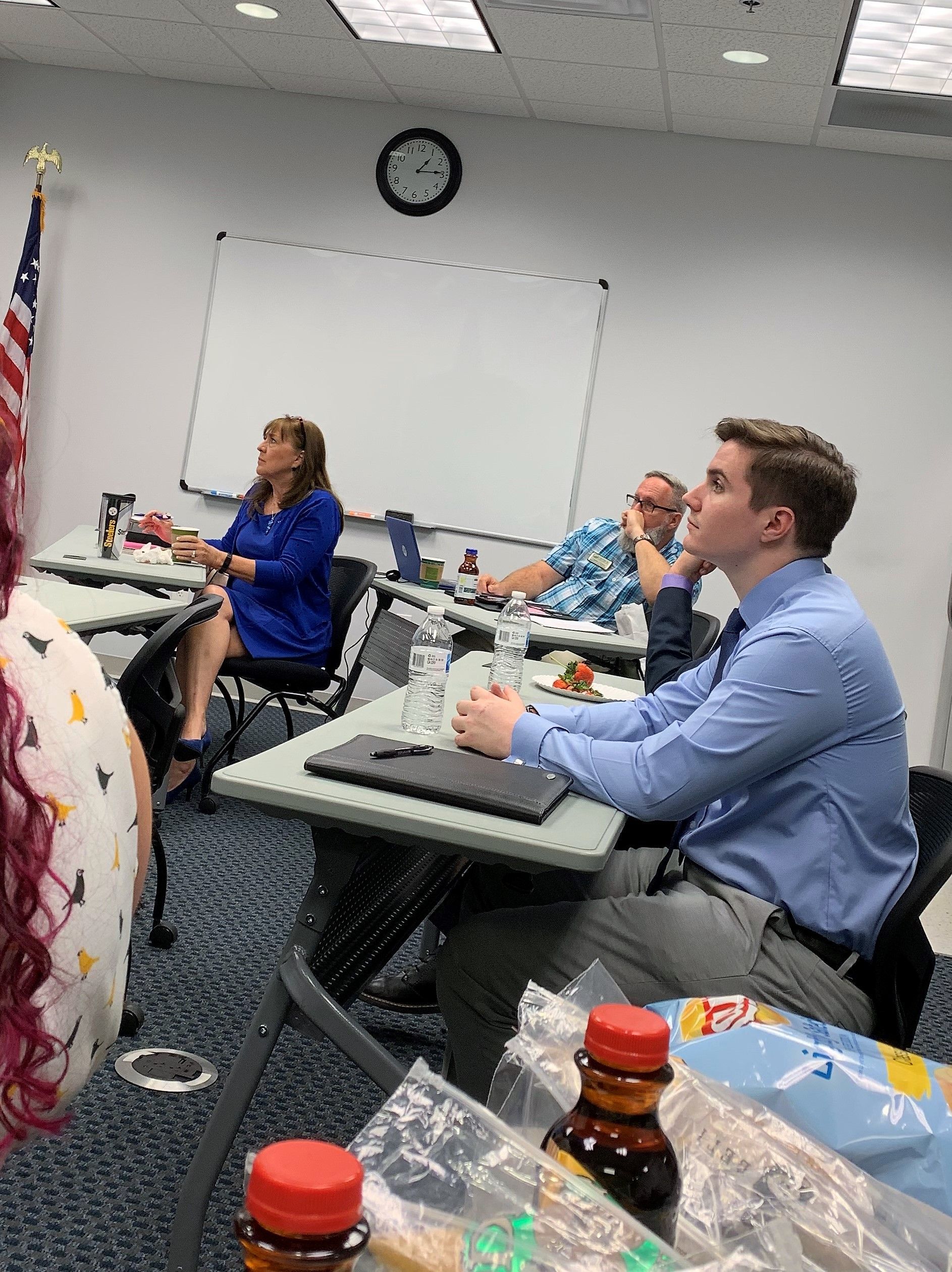 People seated in a classroom. A man in a blue shirt sits at a desk. A woman speaks at the front.