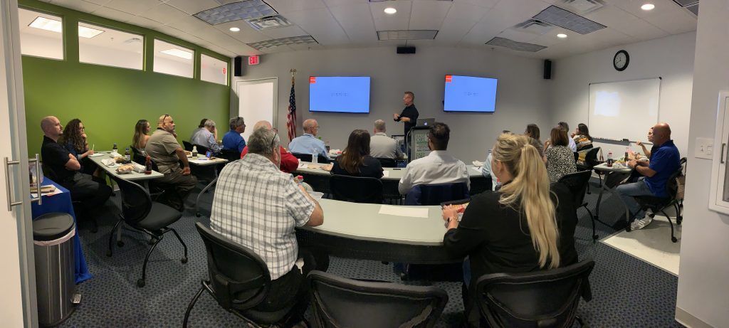 People seated at tables in a conference room, facing a speaker near two screens.