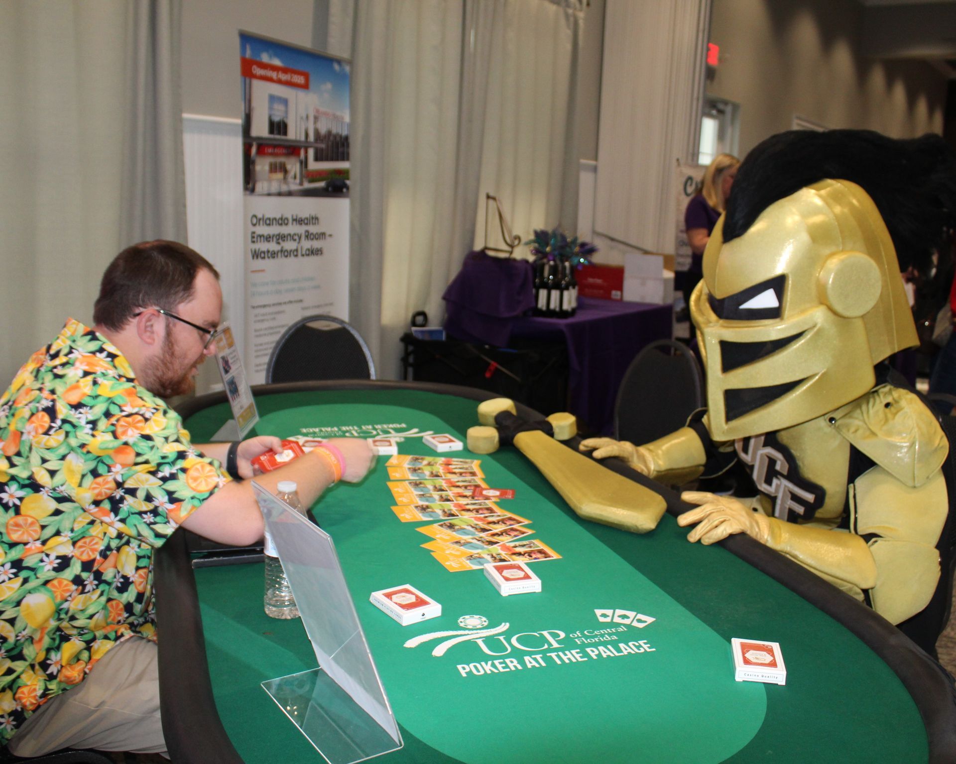 Man in floral shirt and UCF mascot playing poker at a green table with cards and chips.