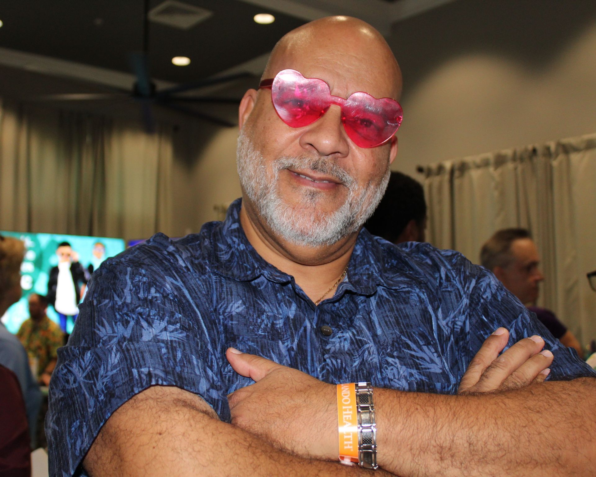 Man wearing heart-shaped pink sunglasses, smiling with arms crossed. Wearing a blue patterned shirt.