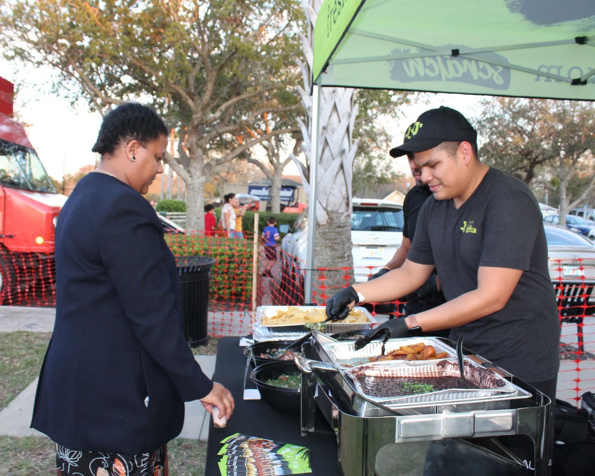 A food vendor serves a customer at an outdoor event. They stand near black food warmer trays, under a green tent.