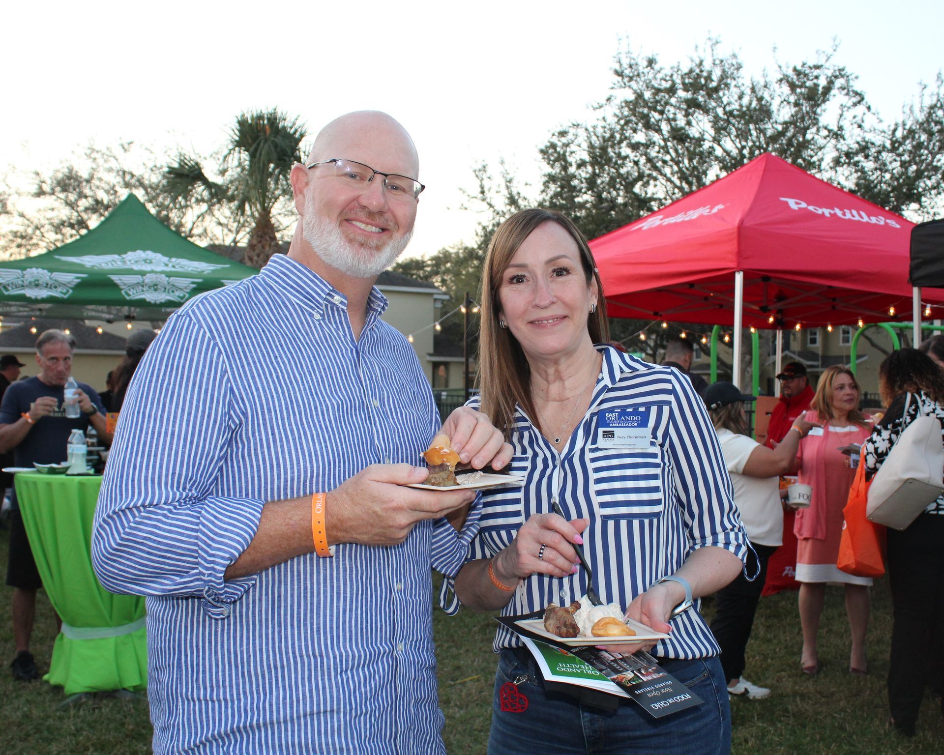 Two people at an outdoor event, holding food, smiling, with tents and other people in the background.