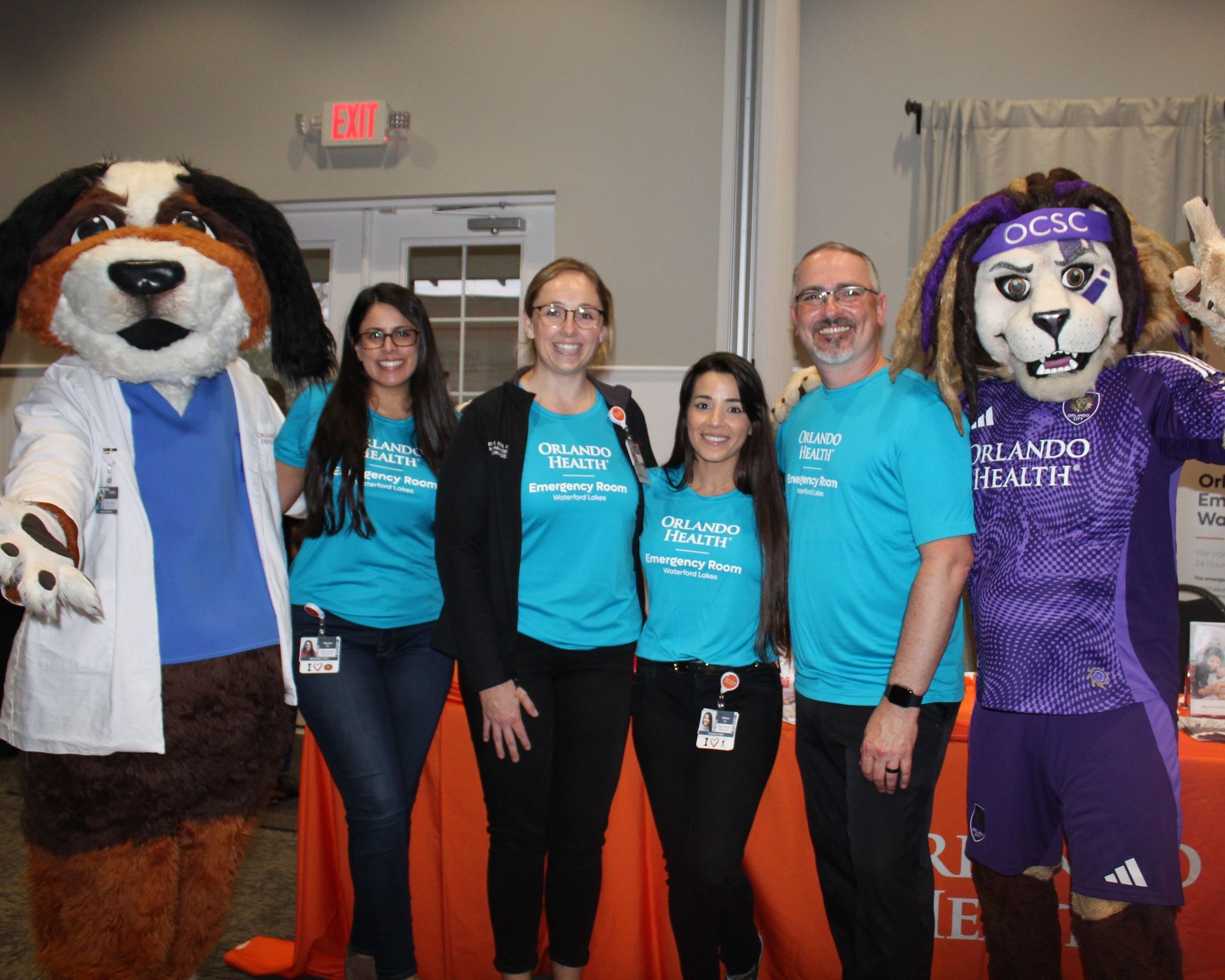 People with mascots at an event. They wear teal shirts with a logo, smiling, and posing for a picture.