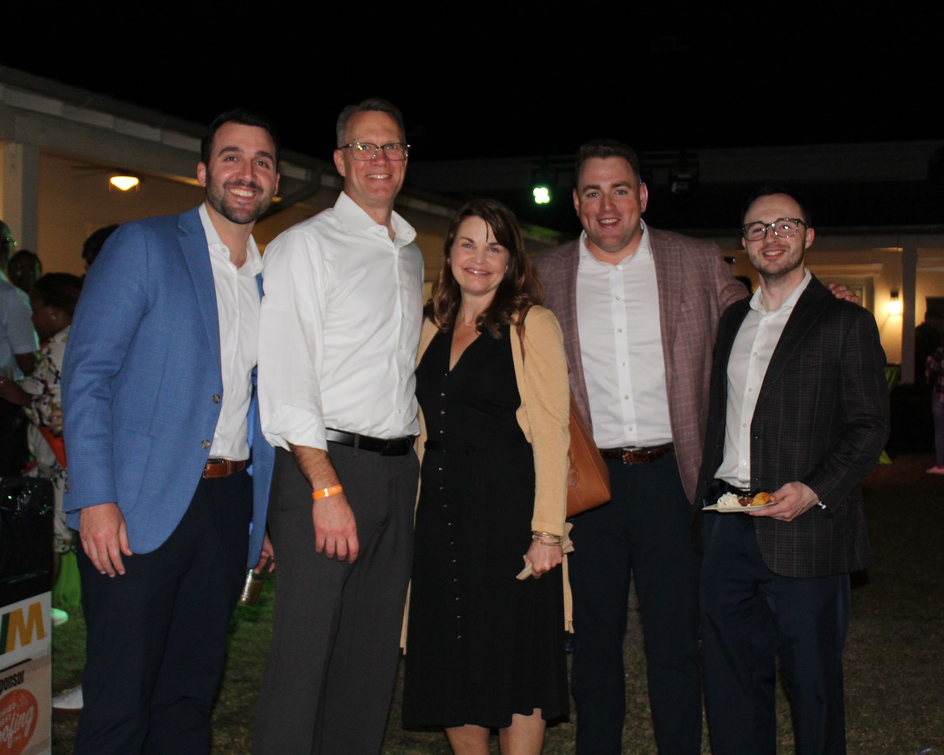 Five people smiling together outdoors, one woman in a black dress, others in blazers, a light building in the background.