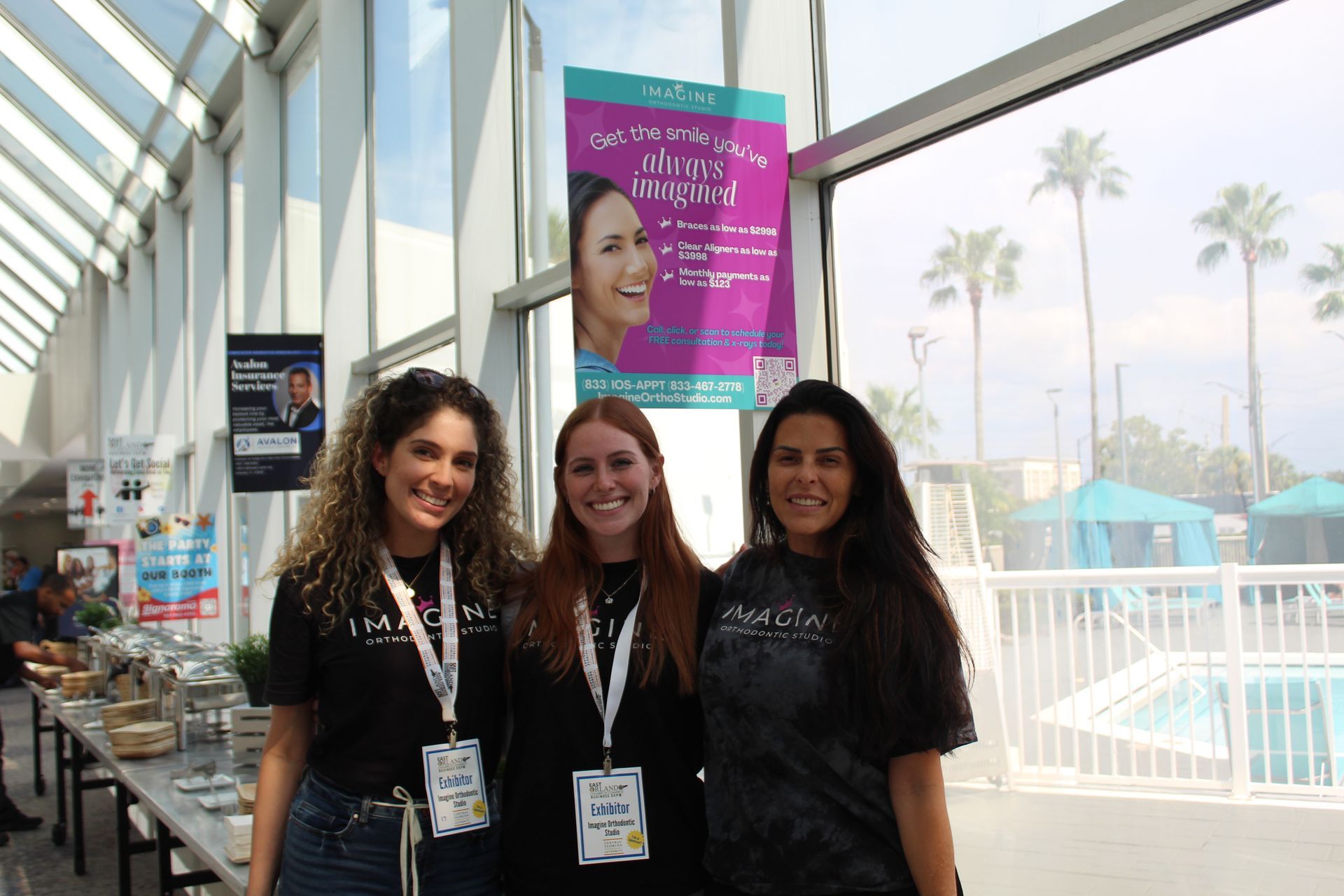 Three women in black shirts stand near a buffet table, posing in front of a window and banner.