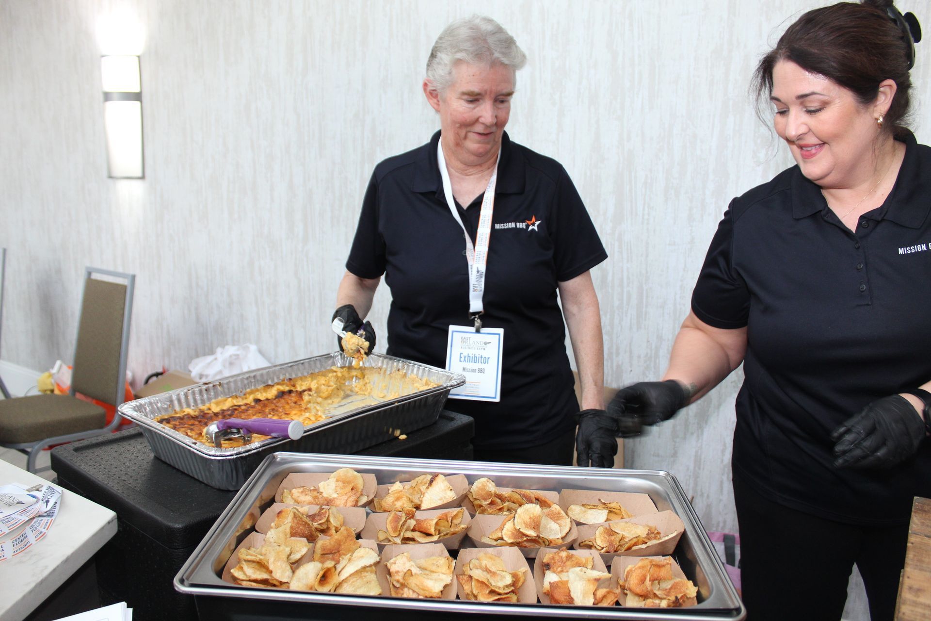 Two people serving food at an event. Woman on the left holds a serving spoon, smiling. Woman on the right, wears gloves.