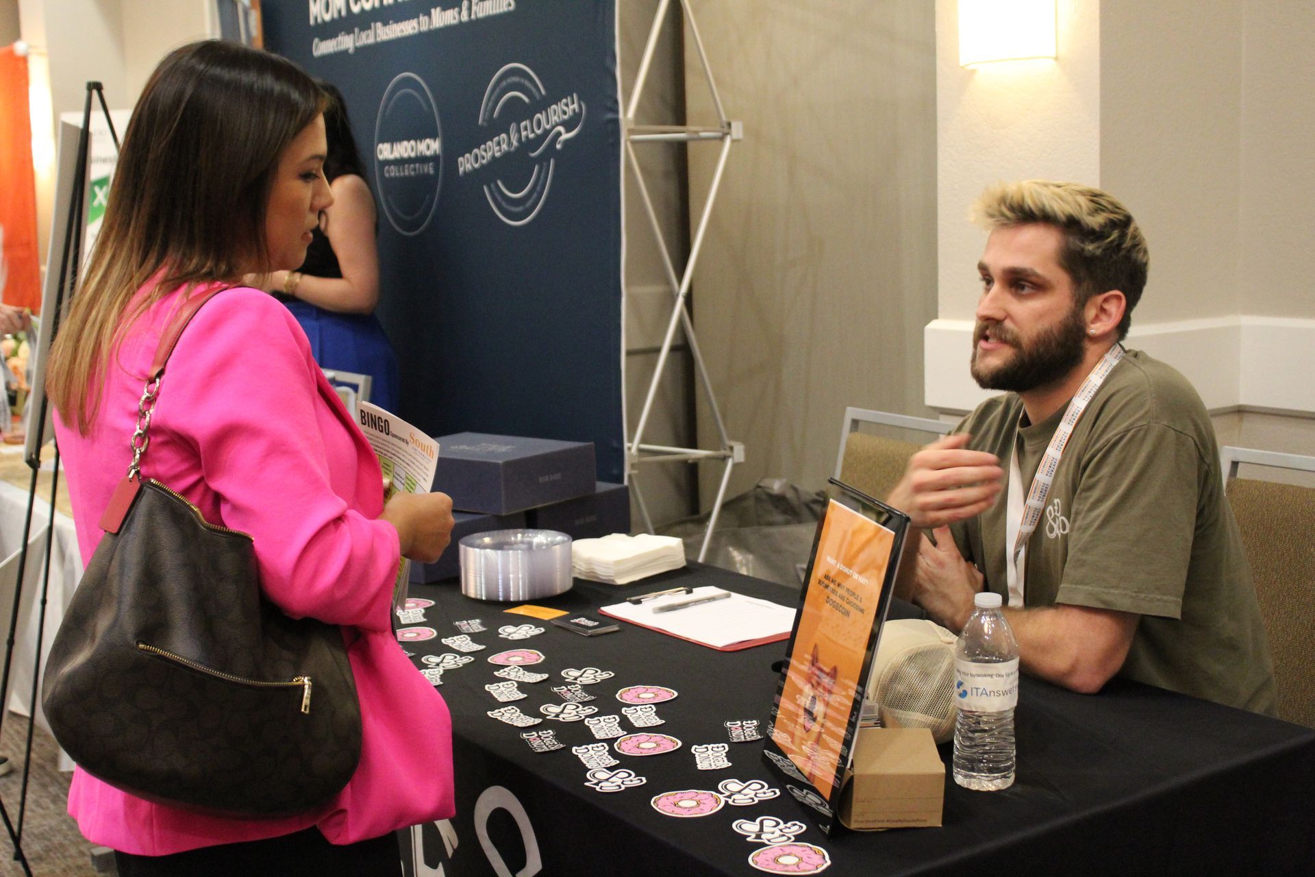 Woman in pink jacket at a table, talking to a man with a beard at a booth.