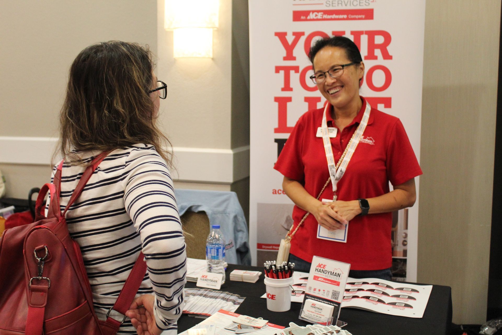 Woman at a booth smiles while talking to another woman; table with brochures and pens; banner.