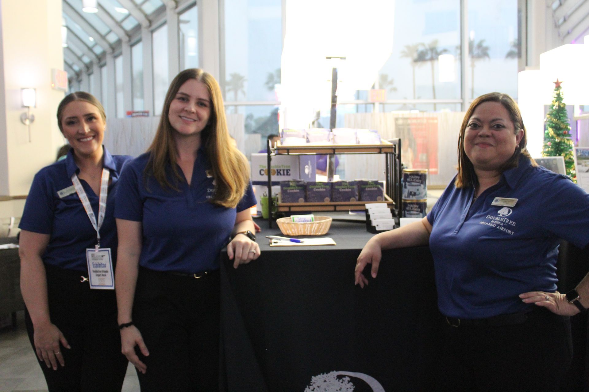 Three women in blue shirts stand behind a table with products at an event.