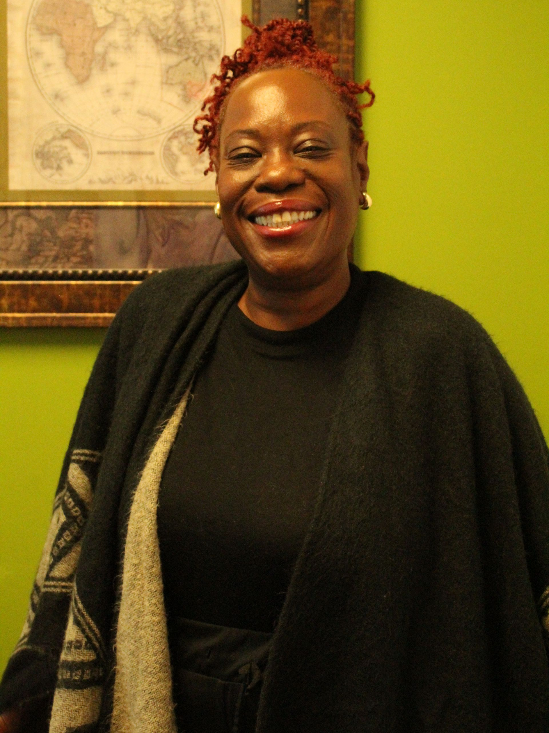 Woman with red-tinted hair smiles. She wears a black top and shawl against a green wall, with a framed map behind her.