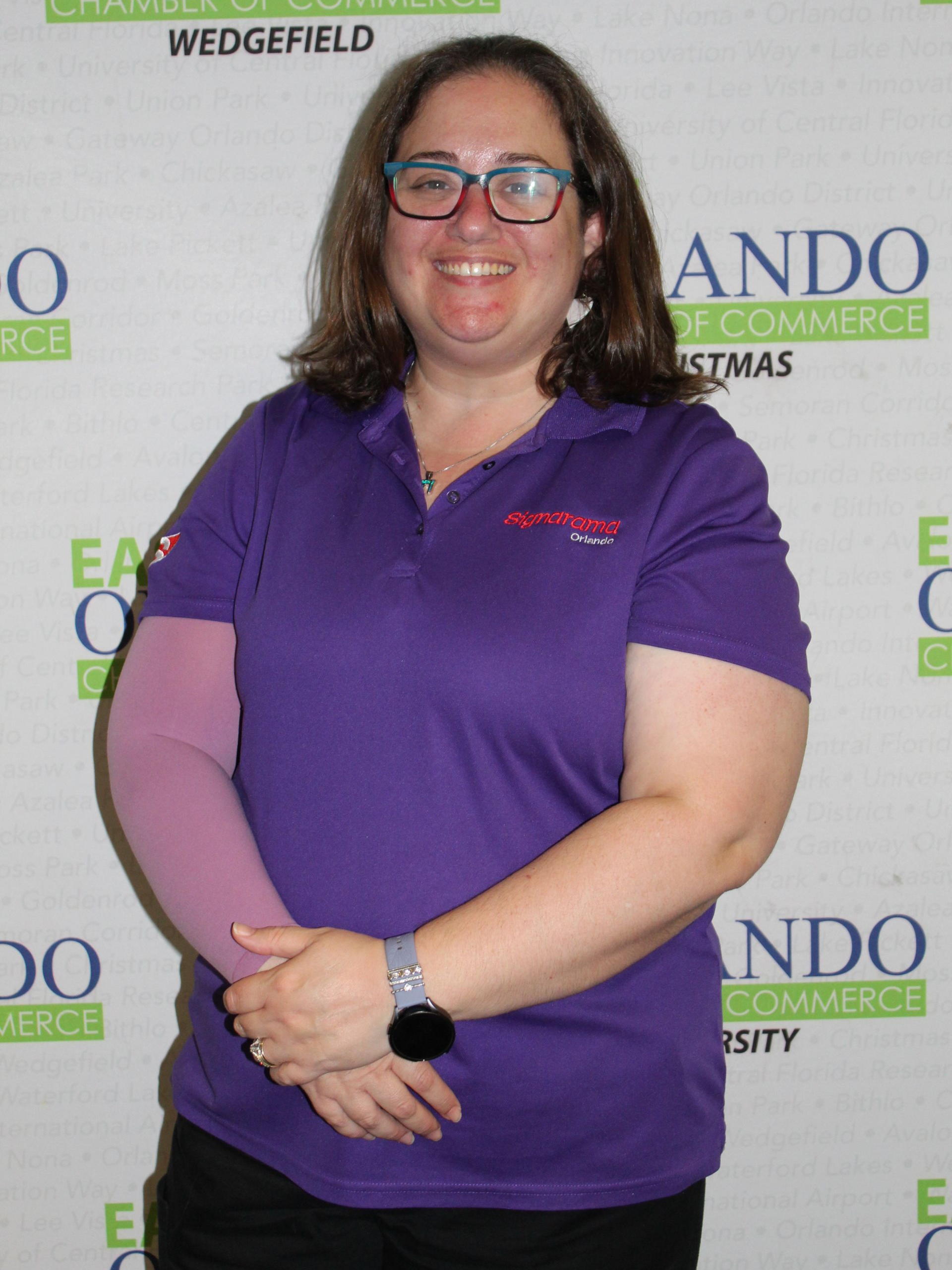 Woman in purple shirt and glasses, wearing a light pink arm sleeve, standing in front of a chamber of commerce backdrop.