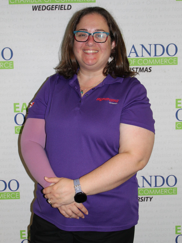 Woman in purple shirt and glasses, wearing a light pink arm sleeve, standing in front of a chamber of commerce backdrop.