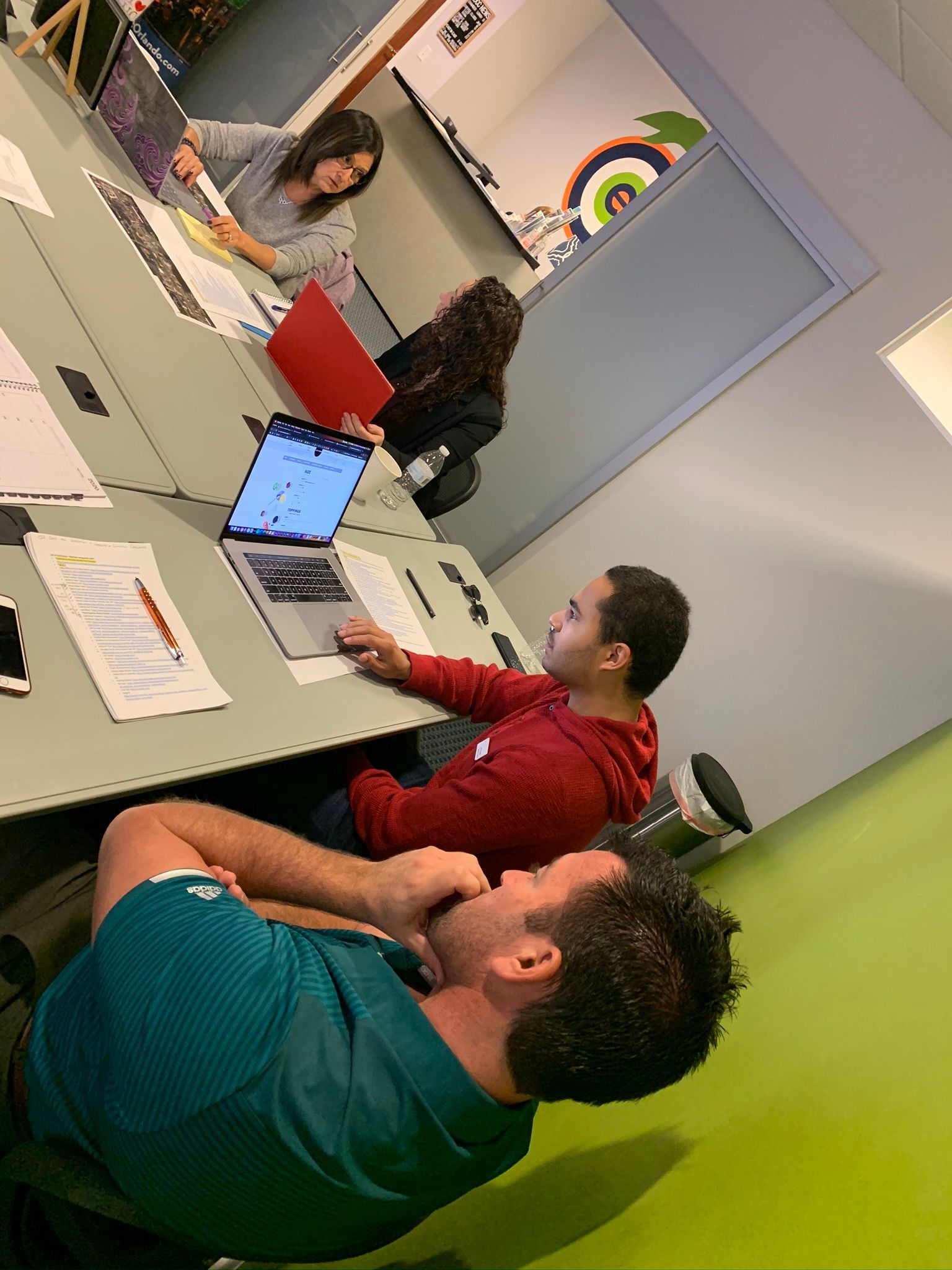 People working at a table with laptops, papers, and a red folder. Inside a room with a green accent wall.