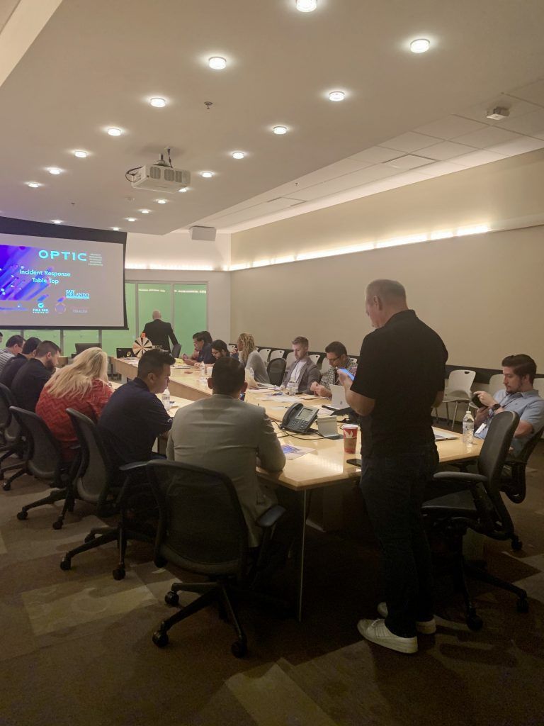 People in a conference room seated around a long table, with a presentation screen and a man standing, speaking.