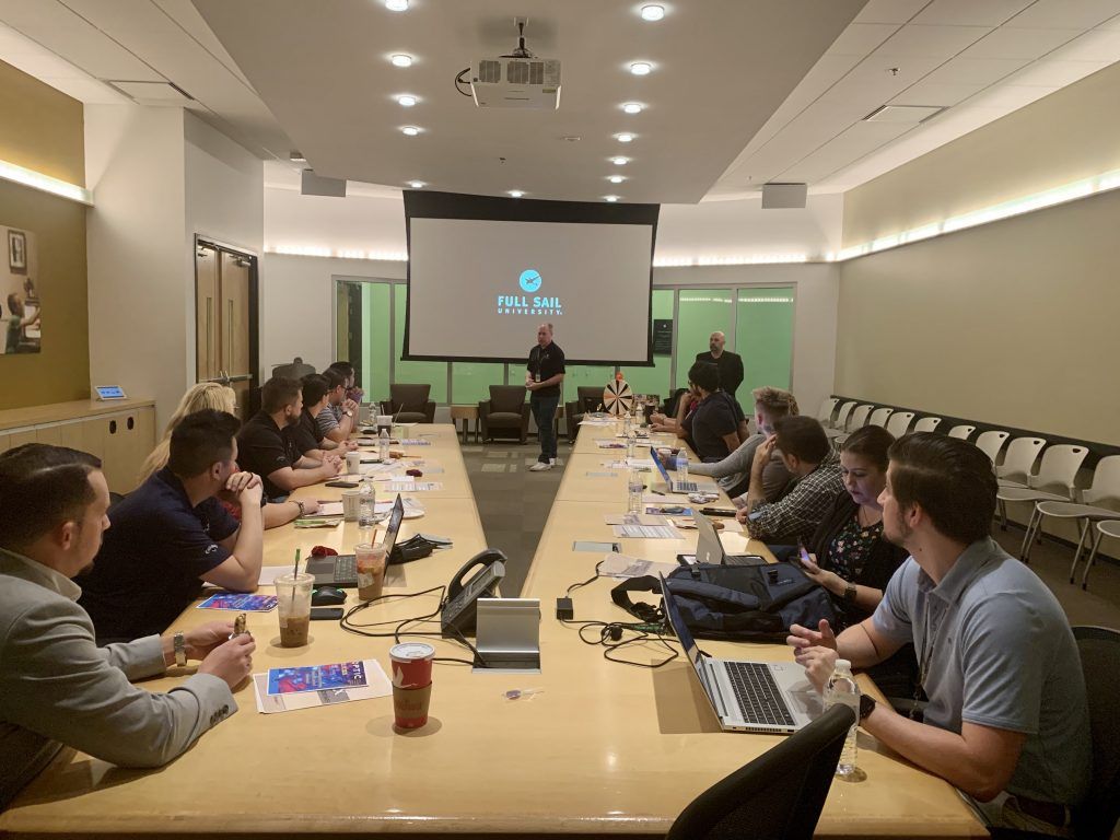 People in a meeting room, listening to a presentation on a large screen with a logo.