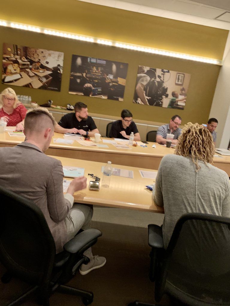 People seated at a table in a meeting, papers and water bottles present. Photographs hang on the wall.