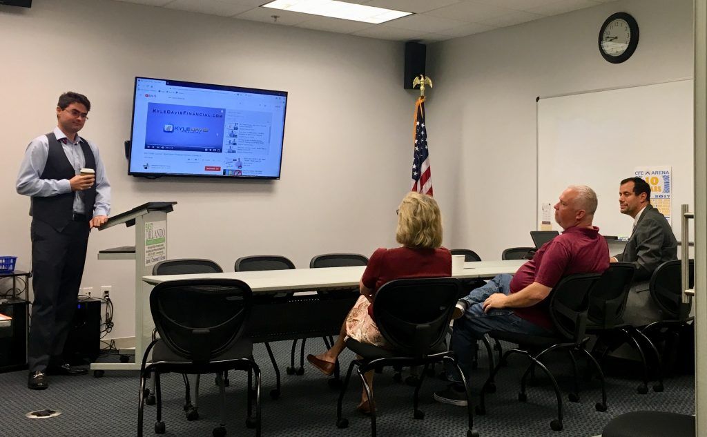 Man presenting in a conference room with three seated listeners and a screen showing a presentation.
