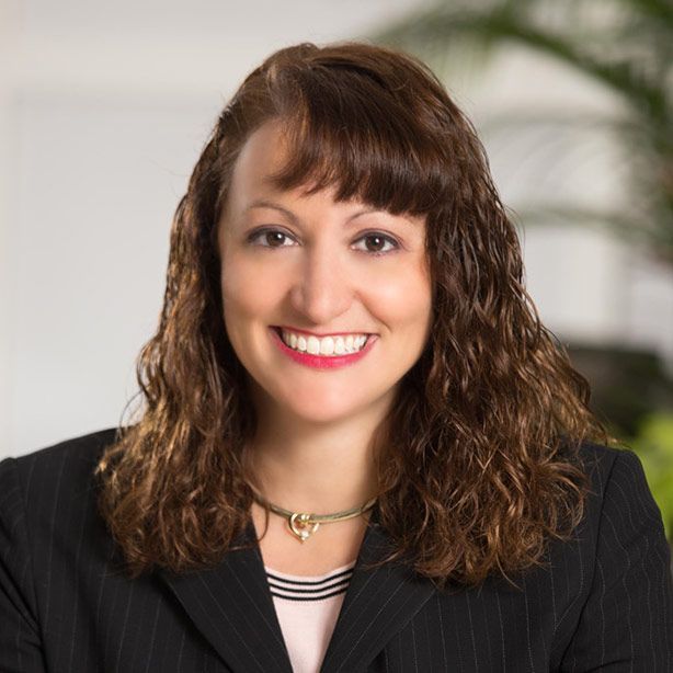 Woman with brown curly hair, wearing a black blazer, smiling, and wearing a gold necklace.