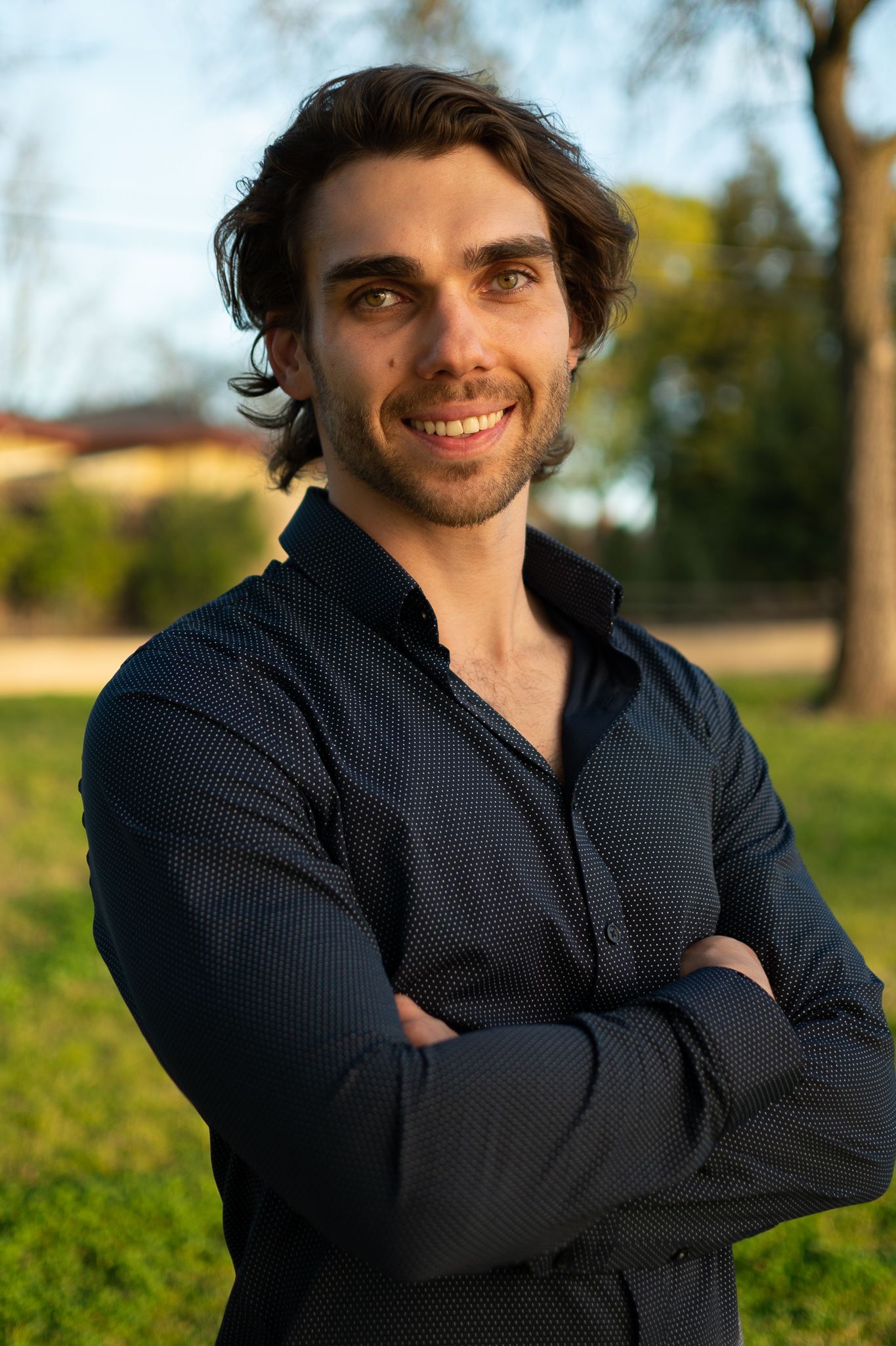 Man with dark curly hair smiles, arms crossed, wearing a dark patterned shirt outdoors.