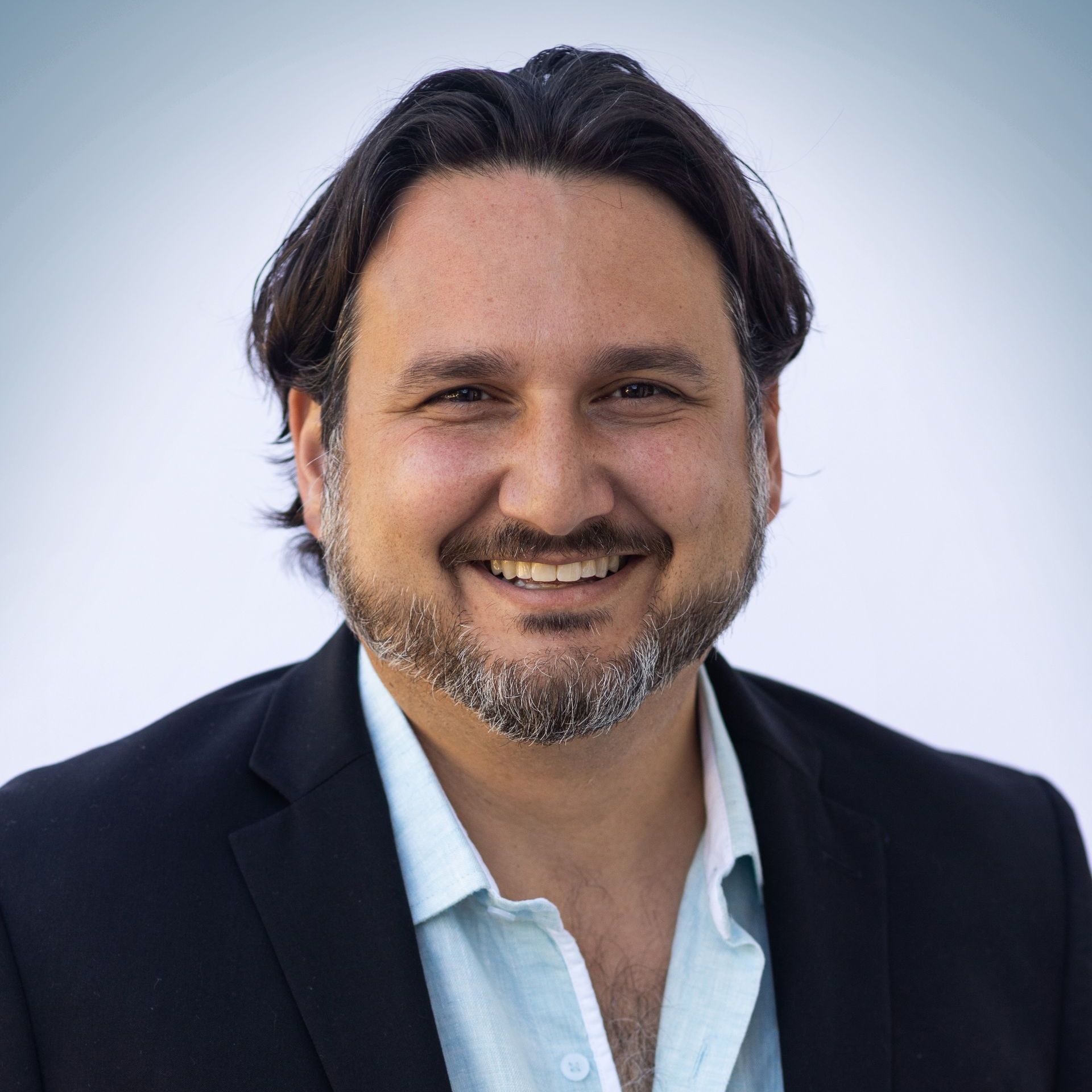 Smiling man in a black blazer and blue shirt, against a gradient background.