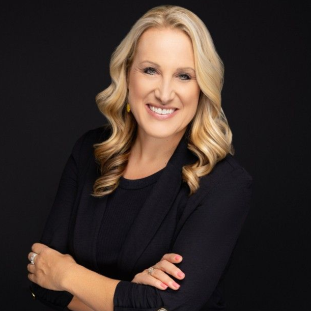 Blonde woman with wavy hair smiles, arms crossed, wearing a dark blazer, against a black backdrop.