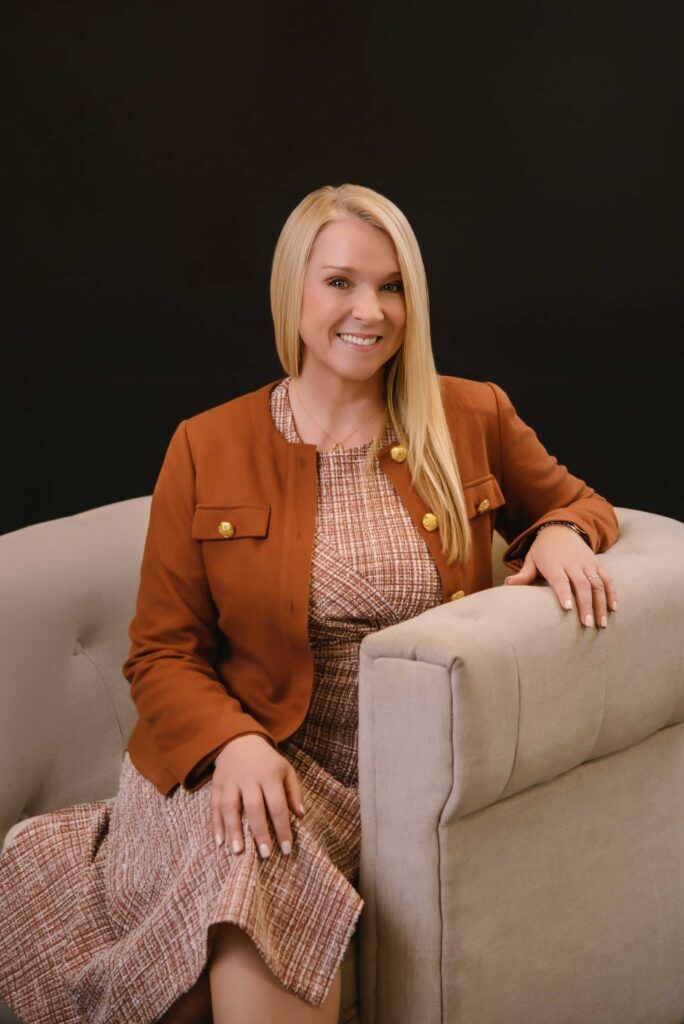 Woman in brown jacket and patterned dress sits on a beige sofa, smiling at the camera. Black background.