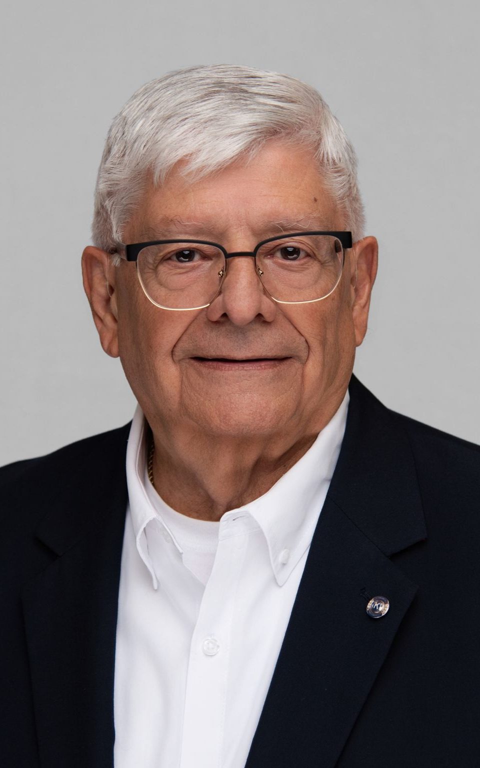 Older man wearing glasses, white shirt, and navy blazer, smiles in front of a neutral background.