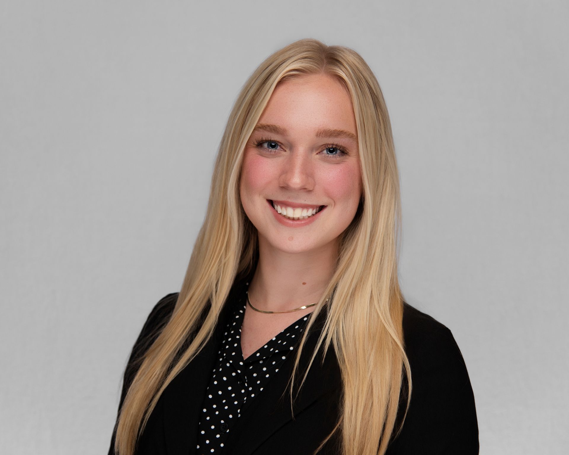 Young woman with long blonde hair, smiling at the camera, wearing a black blazer and patterned top.
