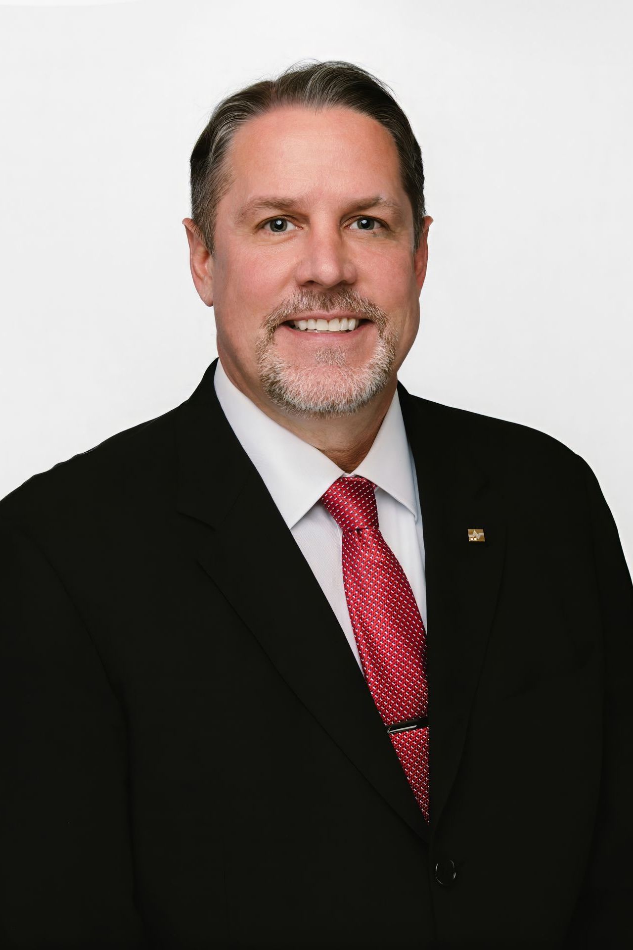 Man in a suit smiles, wearing a red tie. White background.