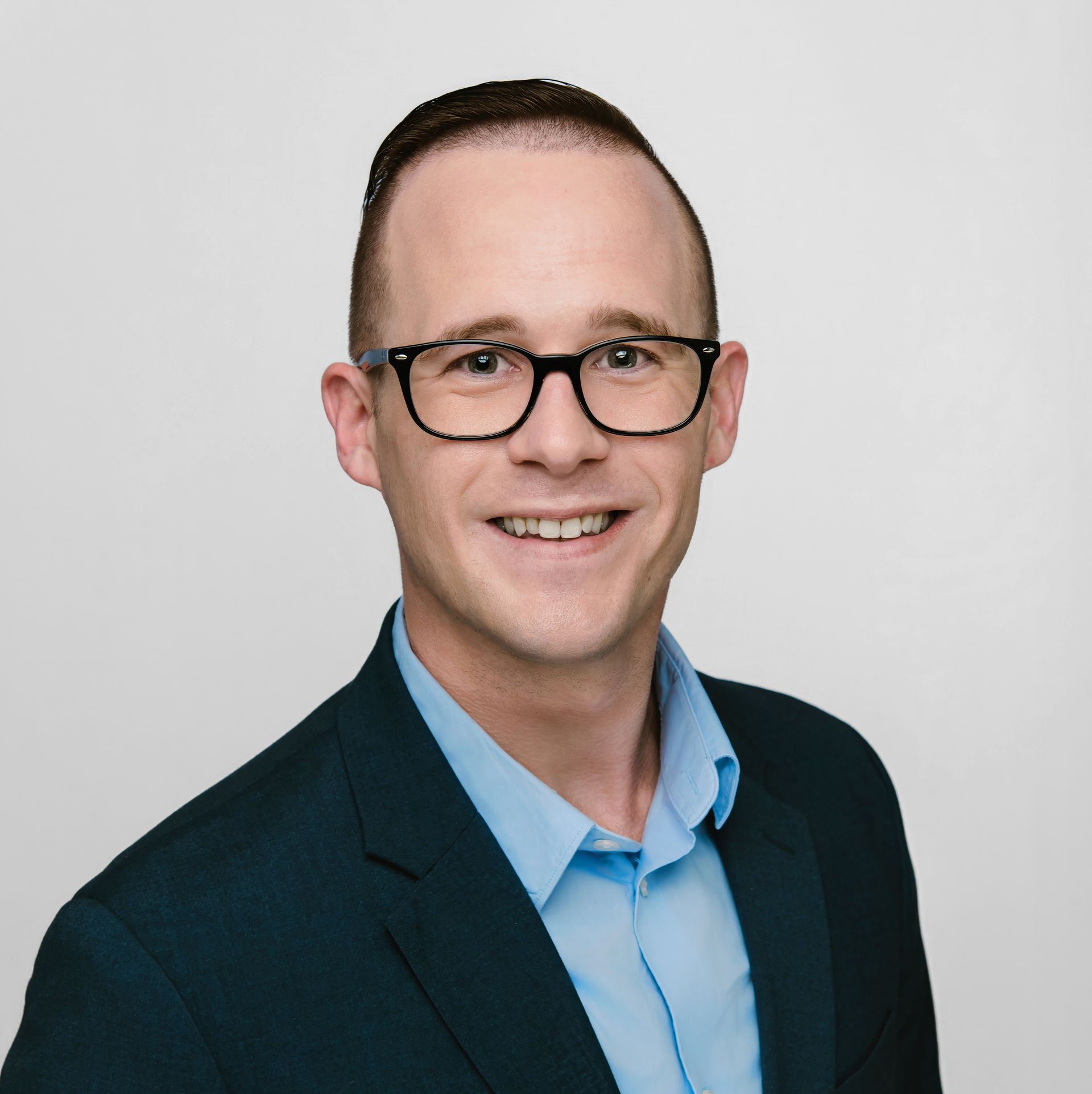 Man with glasses smiling, wearing a blue shirt and dark suit jacket against a light gray background.