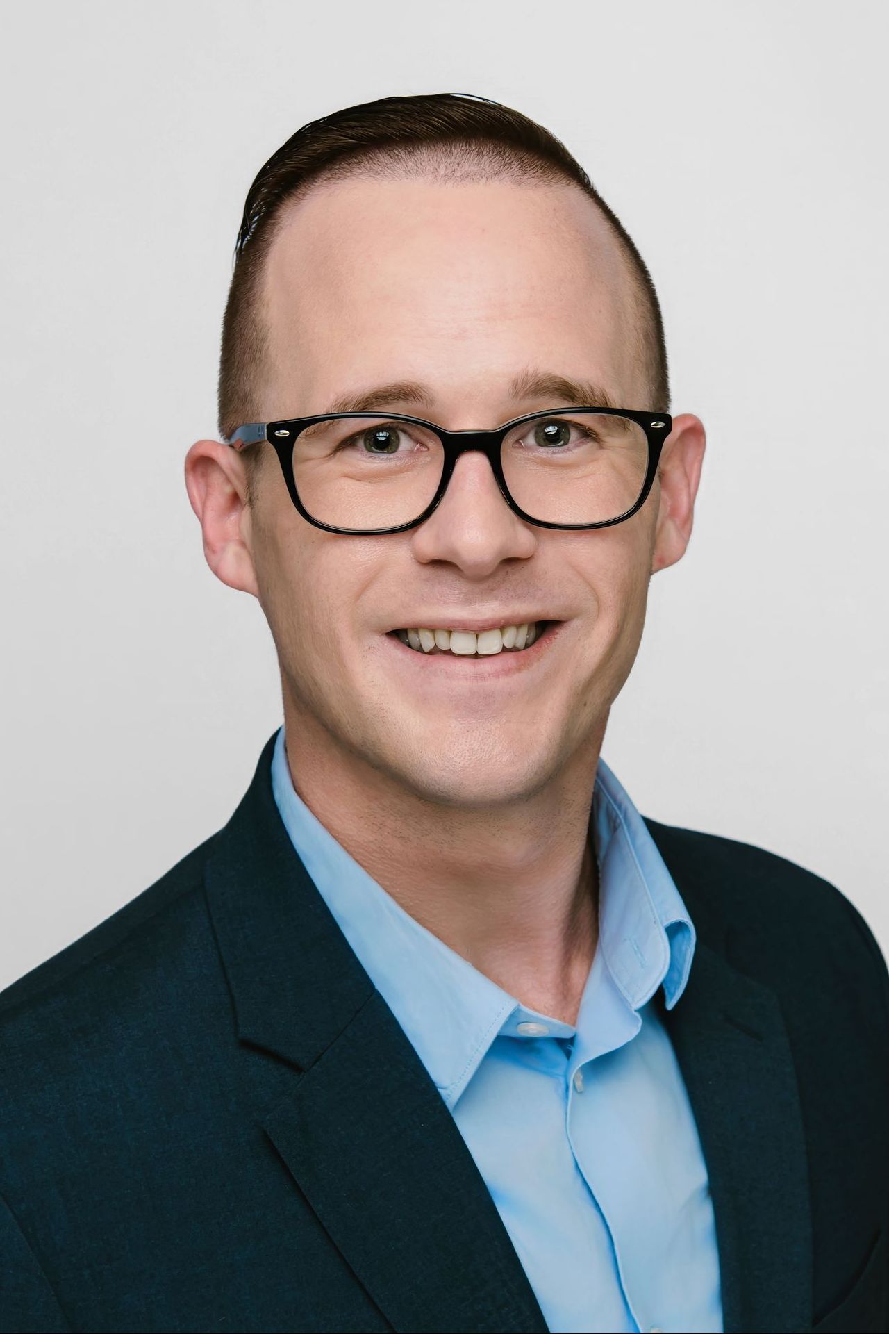 Man with glasses smiles, wearing blue button-down shirt and navy blazer against a neutral background.