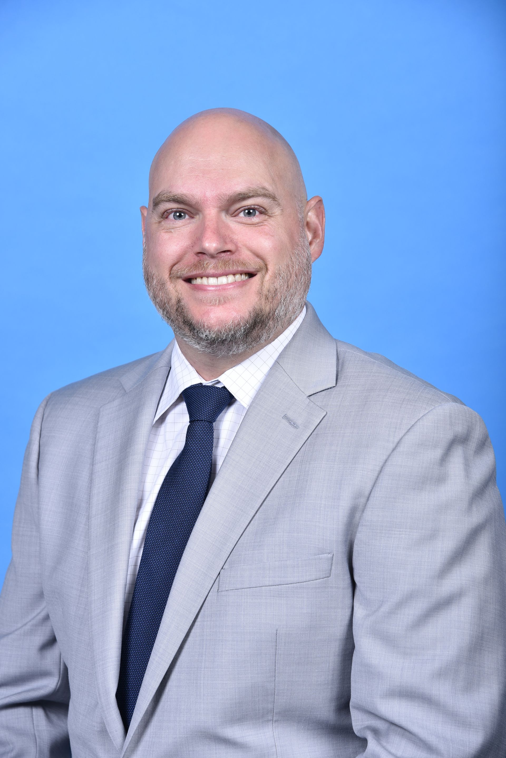 Bald man in gray suit and blue tie smiles at the camera against a blue background.