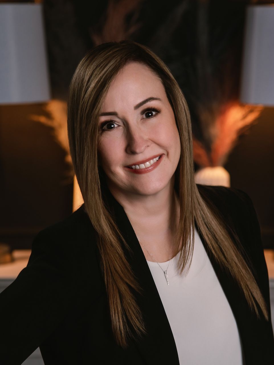 Woman with long, light brown hair smiles, wearing a black blazer and white top; neutral background.