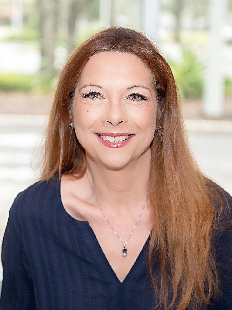 Woman with reddish-brown hair, smiling at the camera, wearing a navy top and silver necklace.