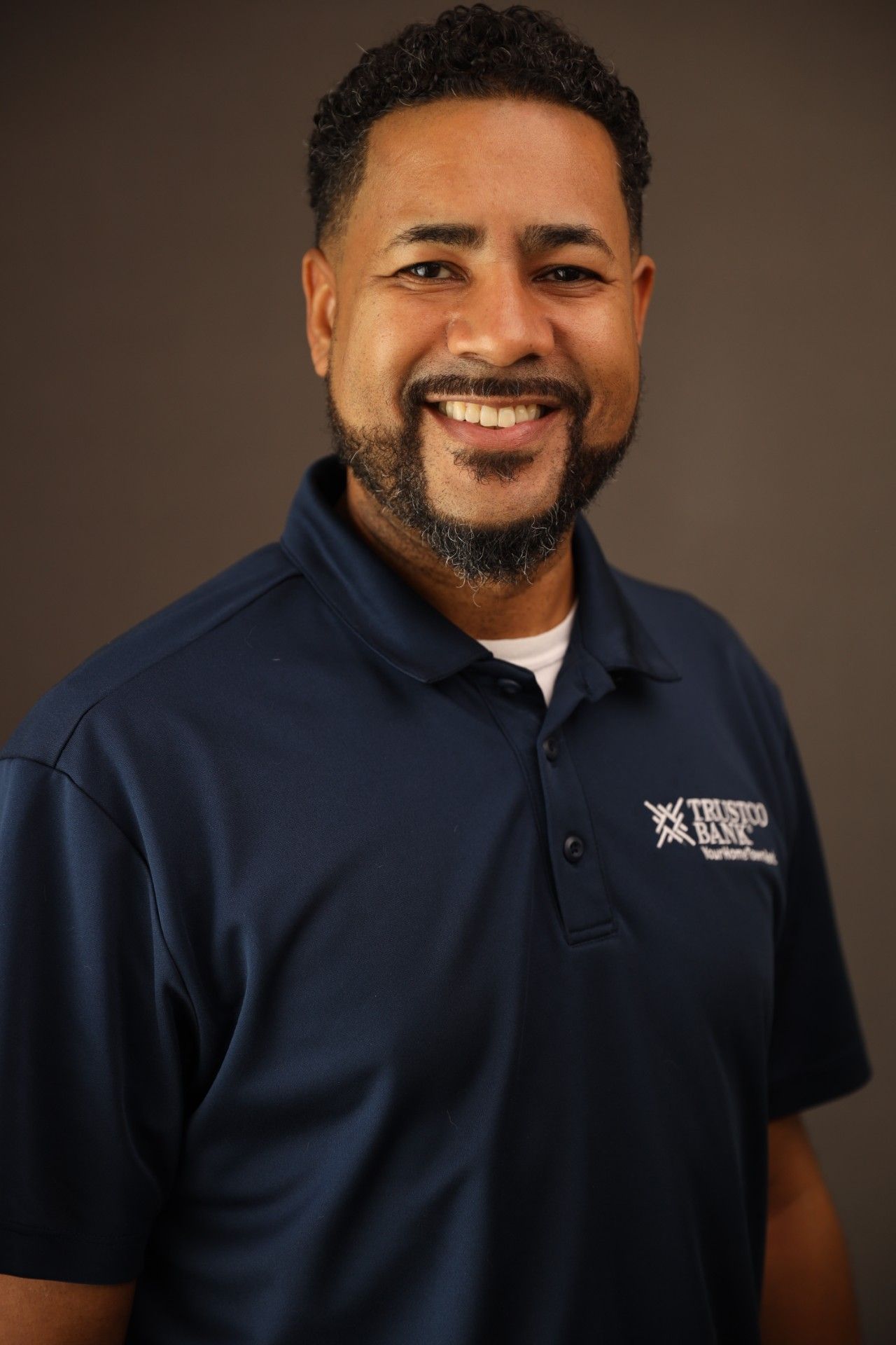 Man smiling, wearing a navy polo shirt with logo, indoors against a gray background.