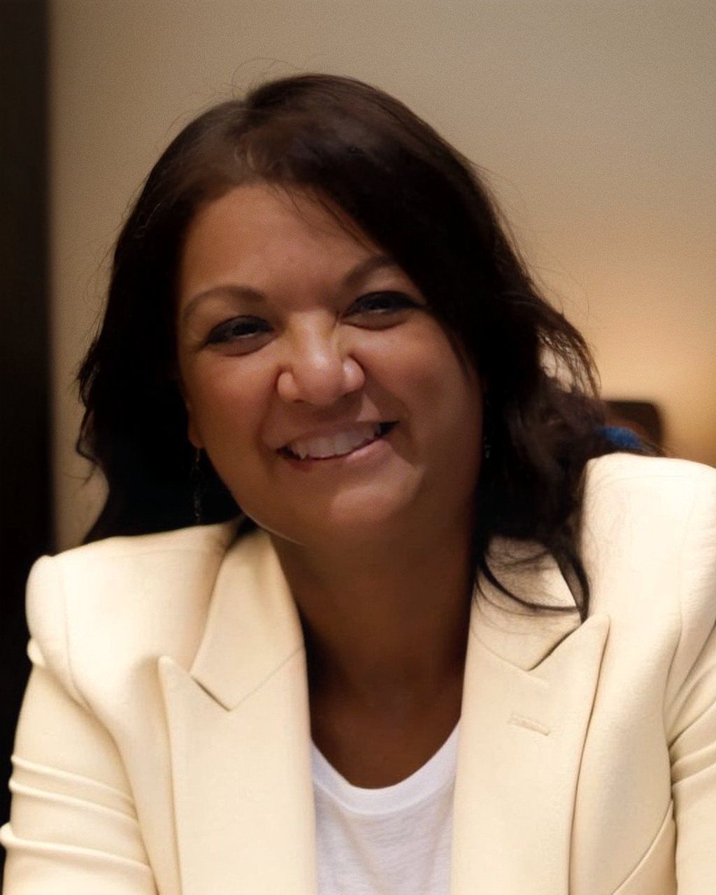 Woman in a cream-colored blazer smiles. Dark hair, white shirt. Indoor setting.