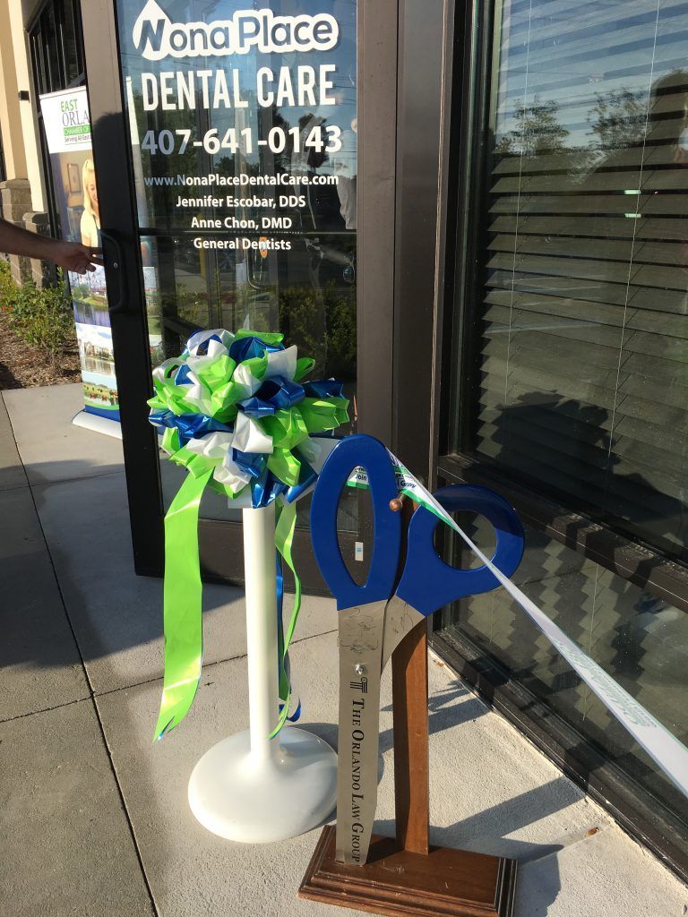 Dental office grand opening ribbon-cutting ceremony with giant blue scissors. Ribbons in green and blue. Door reads 