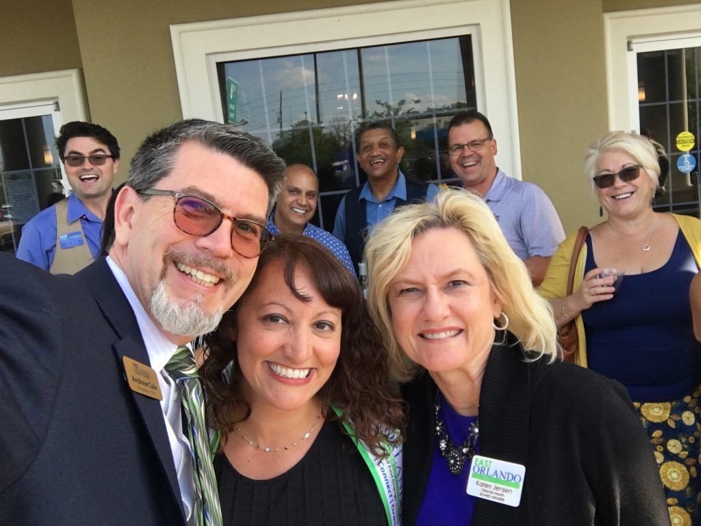 Group smiling for a selfie outside a building. People of various ethnicities wear casual and professional attire.