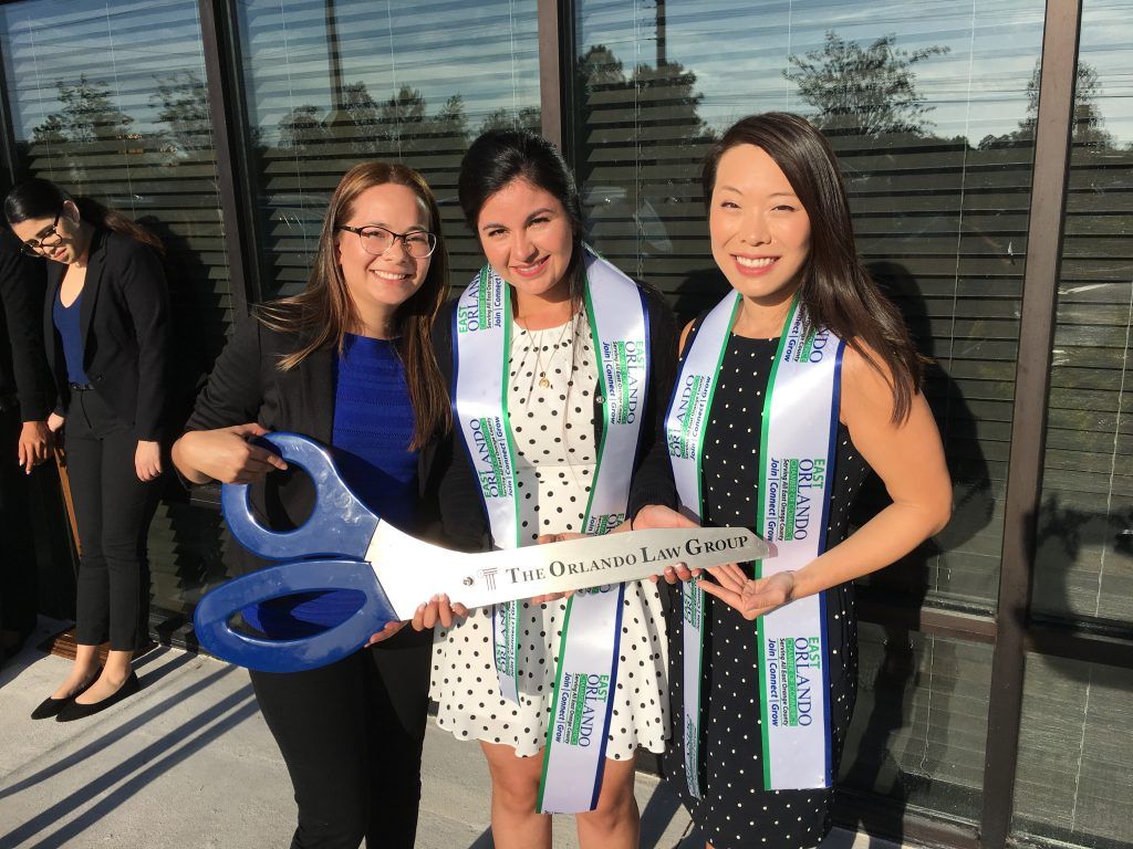 Three women hold a giant pair of scissors and a sign. They wear sashes, smiling in front of a modern building.