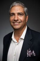 Man in a navy blazer and white shirt smiles at the camera, a pocket square visible.