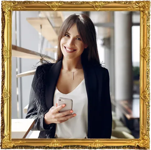 Woman smiling, holding a phone, wearing a black blazer with feather detail, in a building with a wooden staircase, framed.