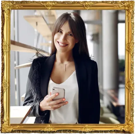 Woman smiling, holding a phone, wearing a black blazer with feather detail, in a building with a wooden staircase, framed.