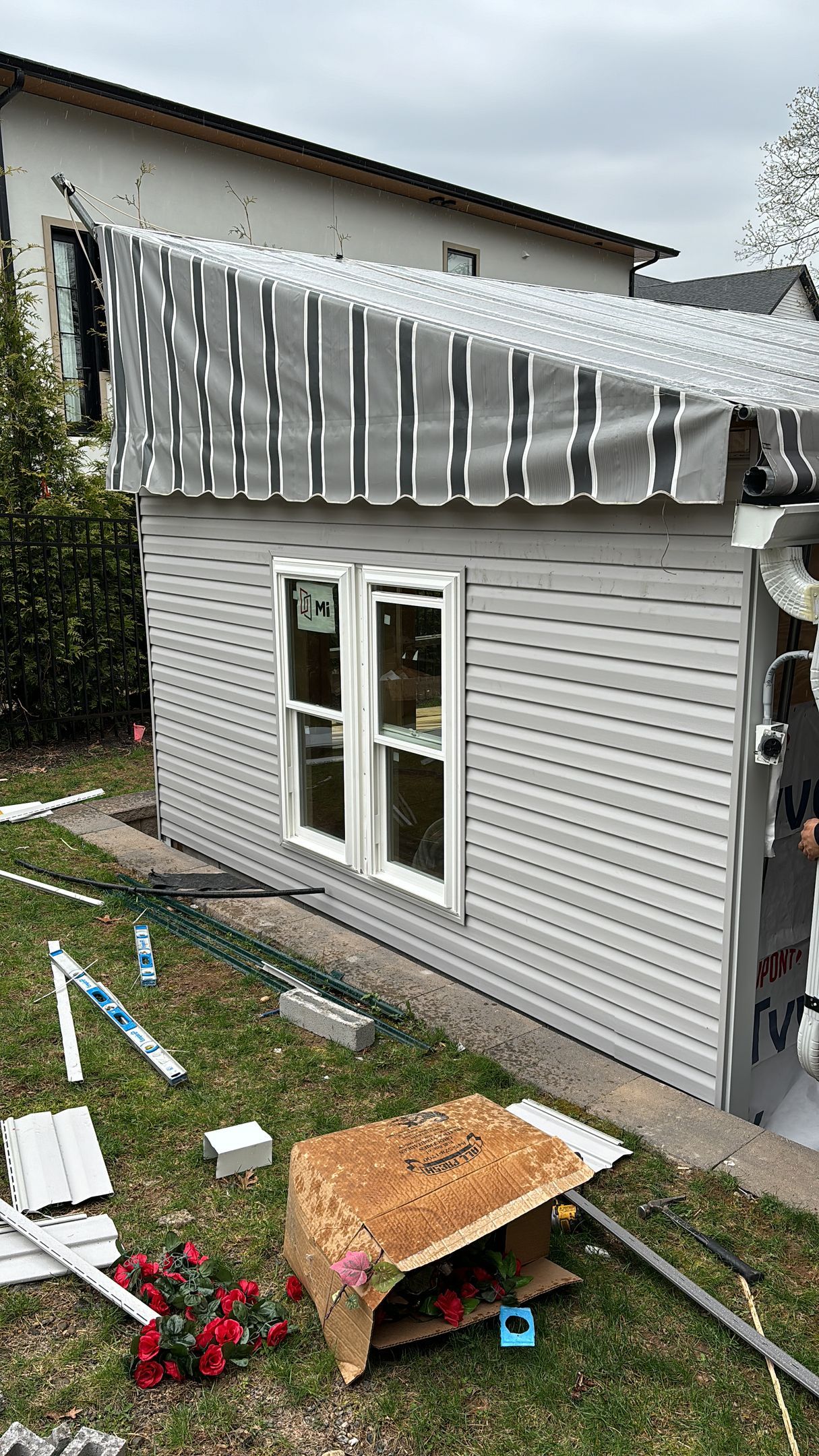 Gray-sided shed with white-framed windows, under construction, in a yard with tools and debris.