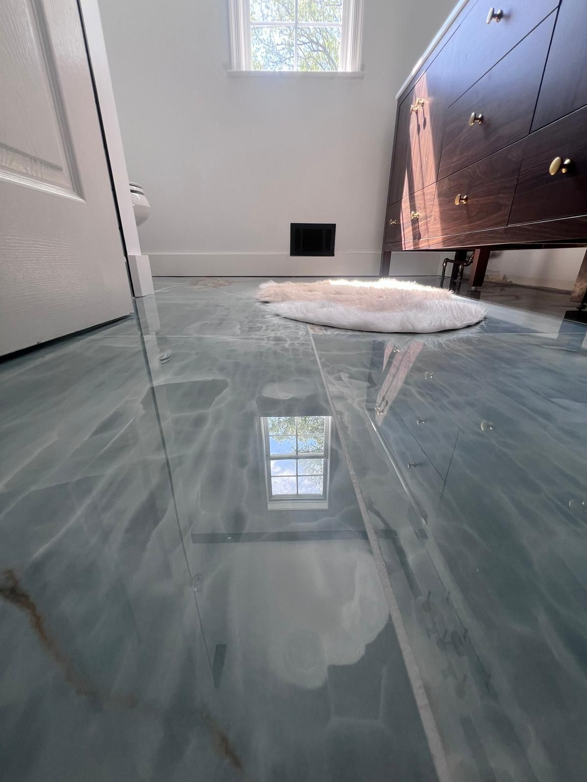 Blue epoxy bathroom floor, window reflected, with a white rug and dark wood cabinet.