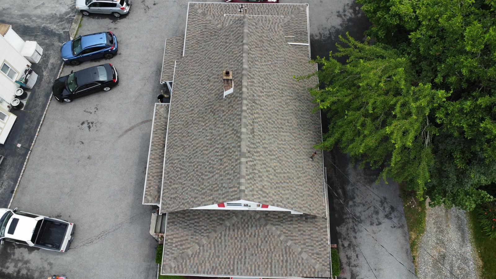 Overhead view of a building with a gray shingled roof, chimney, surrounded by parking lot and green trees.