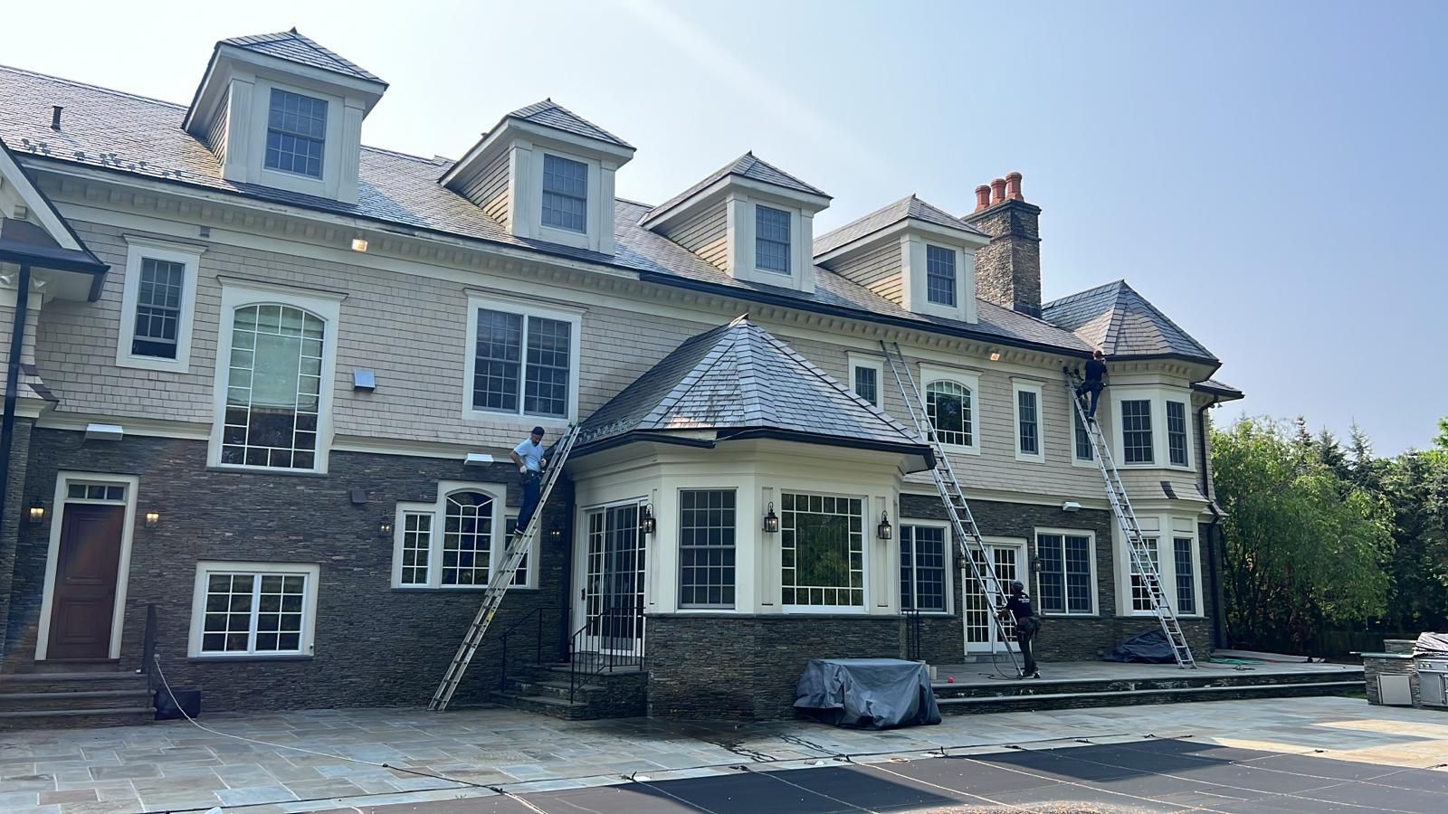 Workers on ladders painting a large beige and brick house with dormer windows on a sunny day.