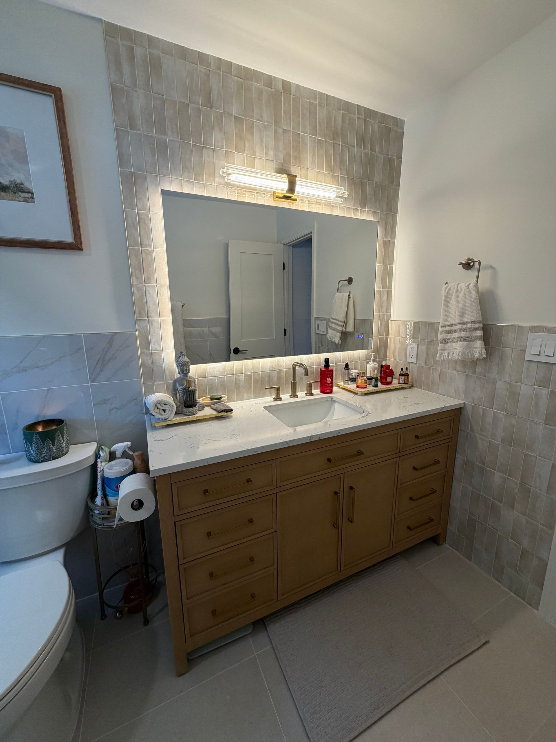 Bathroom with light-colored tile wall, vanity with white countertop, large mirror with lights, and toilet.